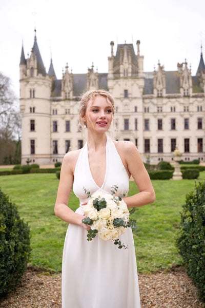 A smiling blonde bride standing against a backdrop of a castle in France wearing a Gaâla halter white bridal silk dress.
