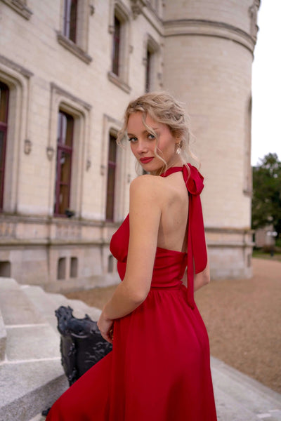 A blonde girl with an elegant hair updo standing in front of a castle in France wearing a vibrant red silk halter Gaâla dress.