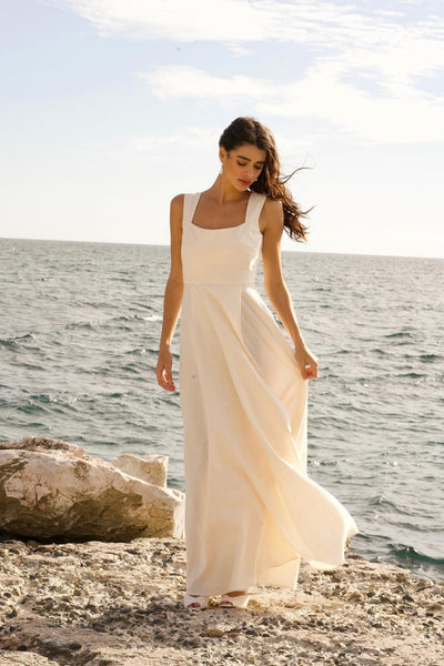 A long haired girl standing in front of the sea wearing a maxi linen wedding dress