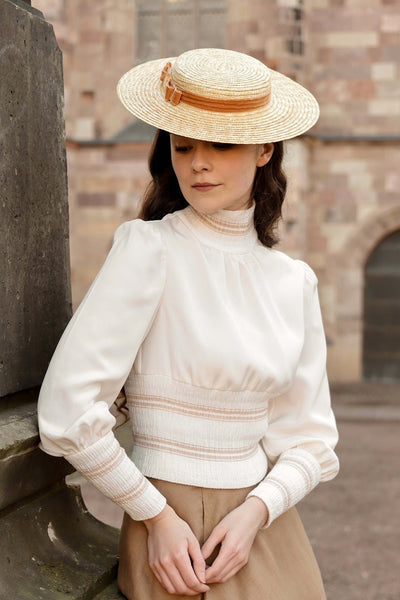 Shirin Altsohn wearing a sun hat while leaning against a wall wearing a Gaâla vintage-inspired high-neck top and beige skirt.