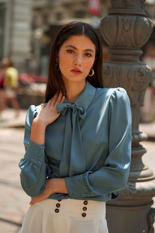 Chic brunette woman in a elegant blue silk blouse with a bow tie neck, leaning against a pillar in Milan.