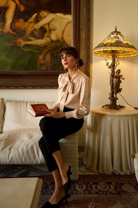 Elegant woman sitting on the arm of a couch in a house with vintage inspired elements and a grand painting, wearing a shiny silk Gaâla blouse with a satin finish