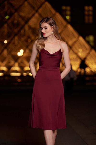 Blonde girl posting in front of the Louvre pyramid in Paris wearing a midi shiny burgundy silk Gaâla dress with sparkly freshwater pearls