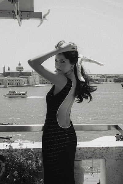 Paola Cossentino standing by the sea in Italy with boats sailing in the background wearing an open back Gaâla maxi knit dress