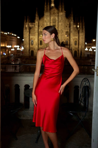 Brunette girl standing on a balcony against a backdrop of the Duomo Cathedral in Milan, wearing a slinky red shiny silk Gaâla cowl-neck dress