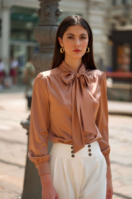 Stylish brunette woman in an elegant champagne-colored Gaâla shiny silk blouse with a charming bow tie neck, complemented by elegant gold hoop earrings, exploring the beauty of Milan