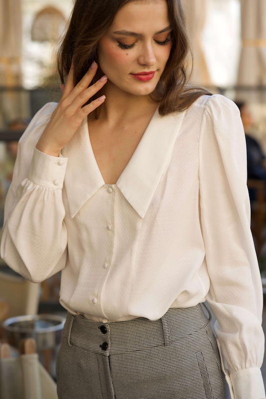 Long-haired smiling woman in Cannes, gently touching her hair while wearing a elegant button-down silk Gaâla blouse with an oversized collar.