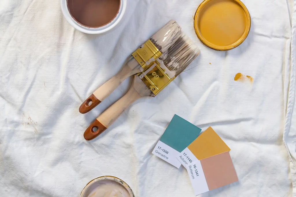 Paint brushes, open paint cans, and pantones on a white cloth covering the floor