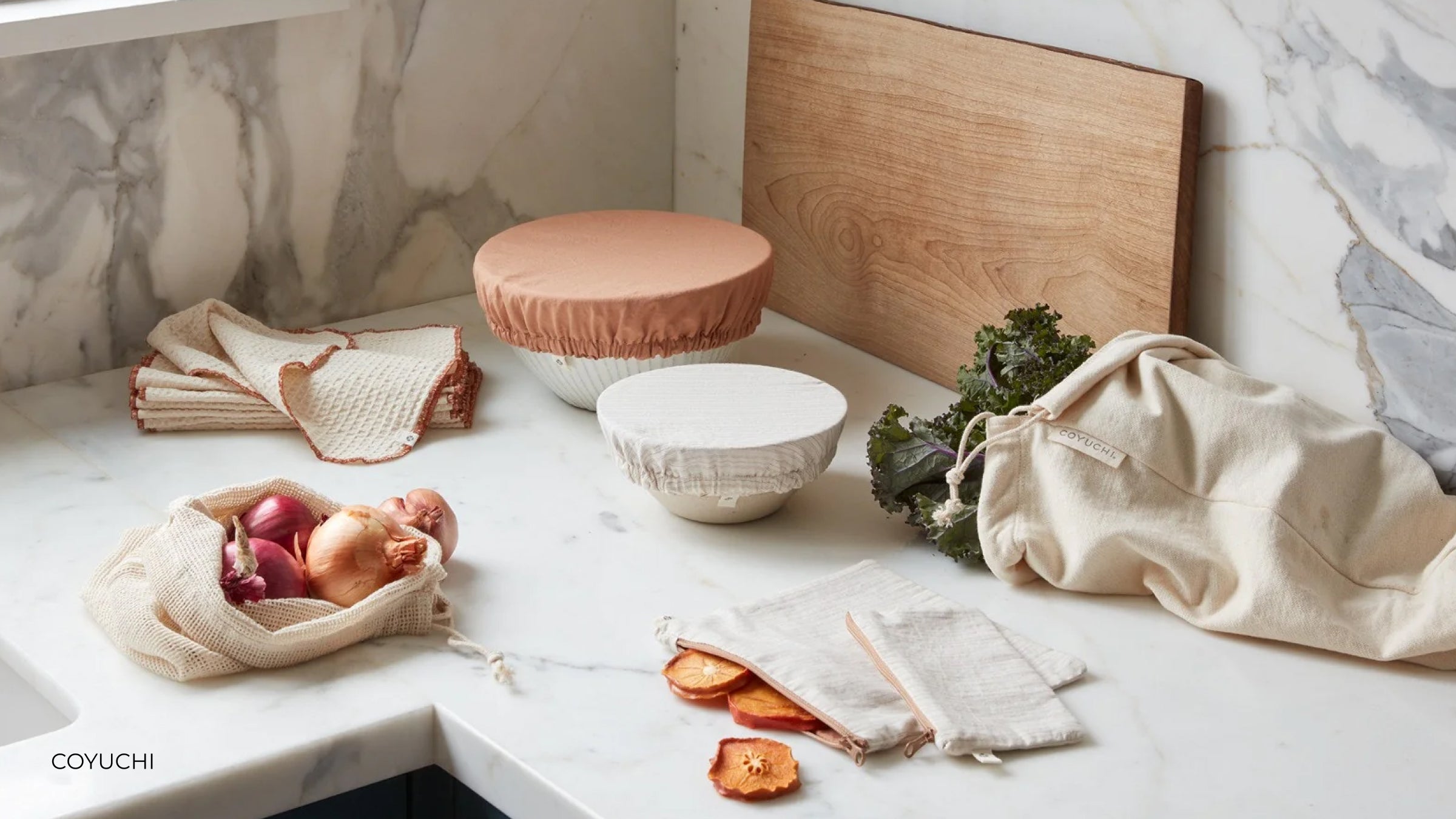 A pile of Coyuchi organic cotton dishcloths sit on a marble countertop with other cotton goods and a wood cutting board