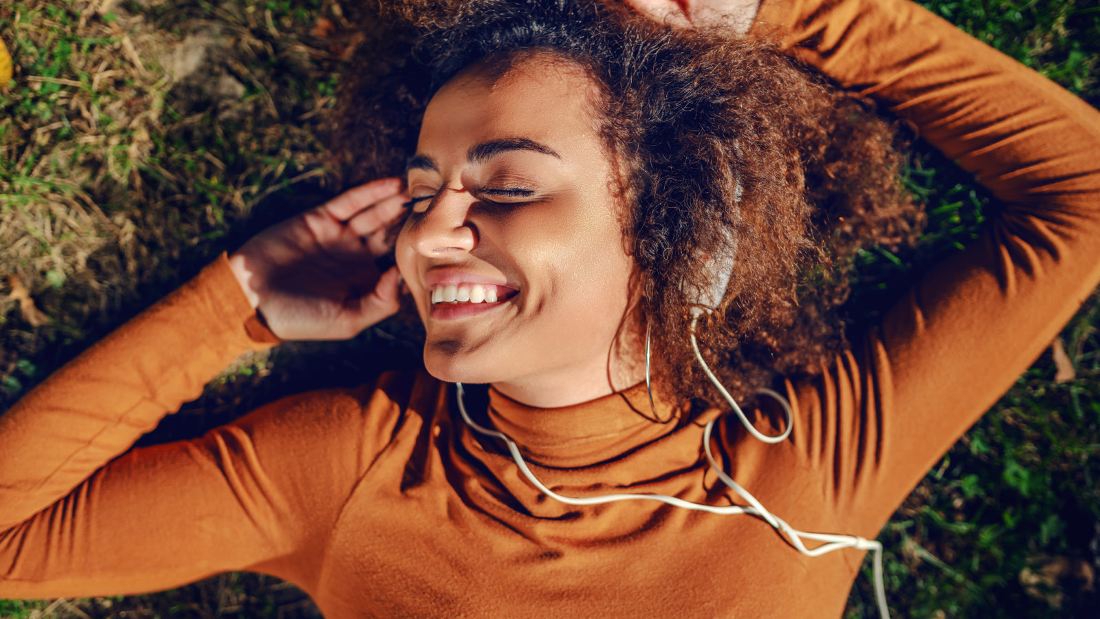A woman with brown curly hair wearing a burnt orange long sleeve shirt listens to headphones while laying on the grass