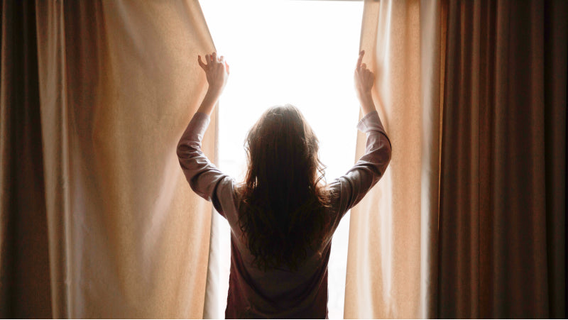 A woman with long brown hair pulls brown curtains shut during the day