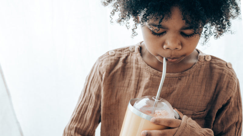 A young black child in brown linen shirt enjoys a delicious morning smoothie in a peach thermos with a stainless steel straw. 