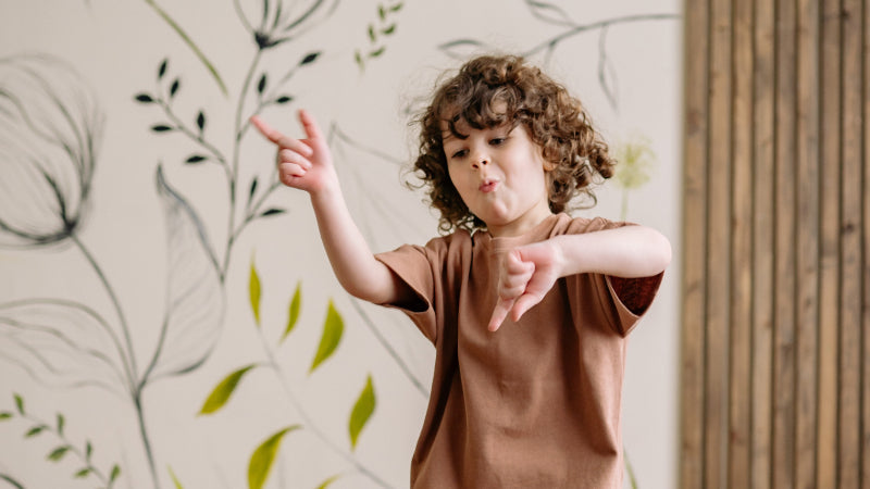 A young child dances as he dresses and puts on a brown shirt in front of wallpaper with a nature motif