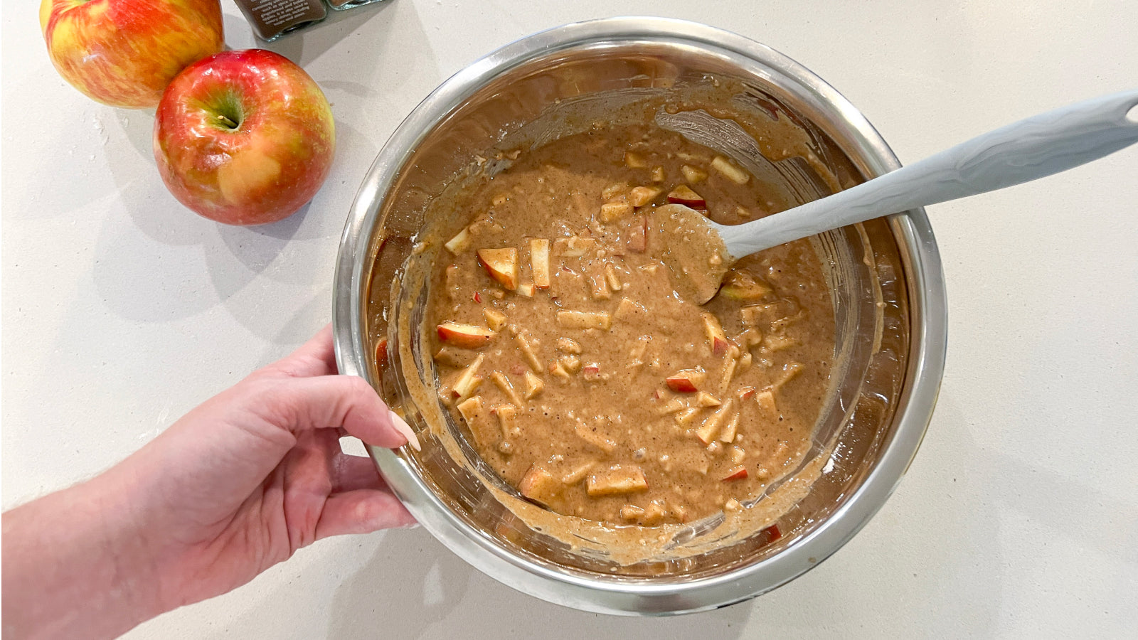 Apple muffin batter mix in a silver bowl with a gray spoon resting on the side and apples on the counter