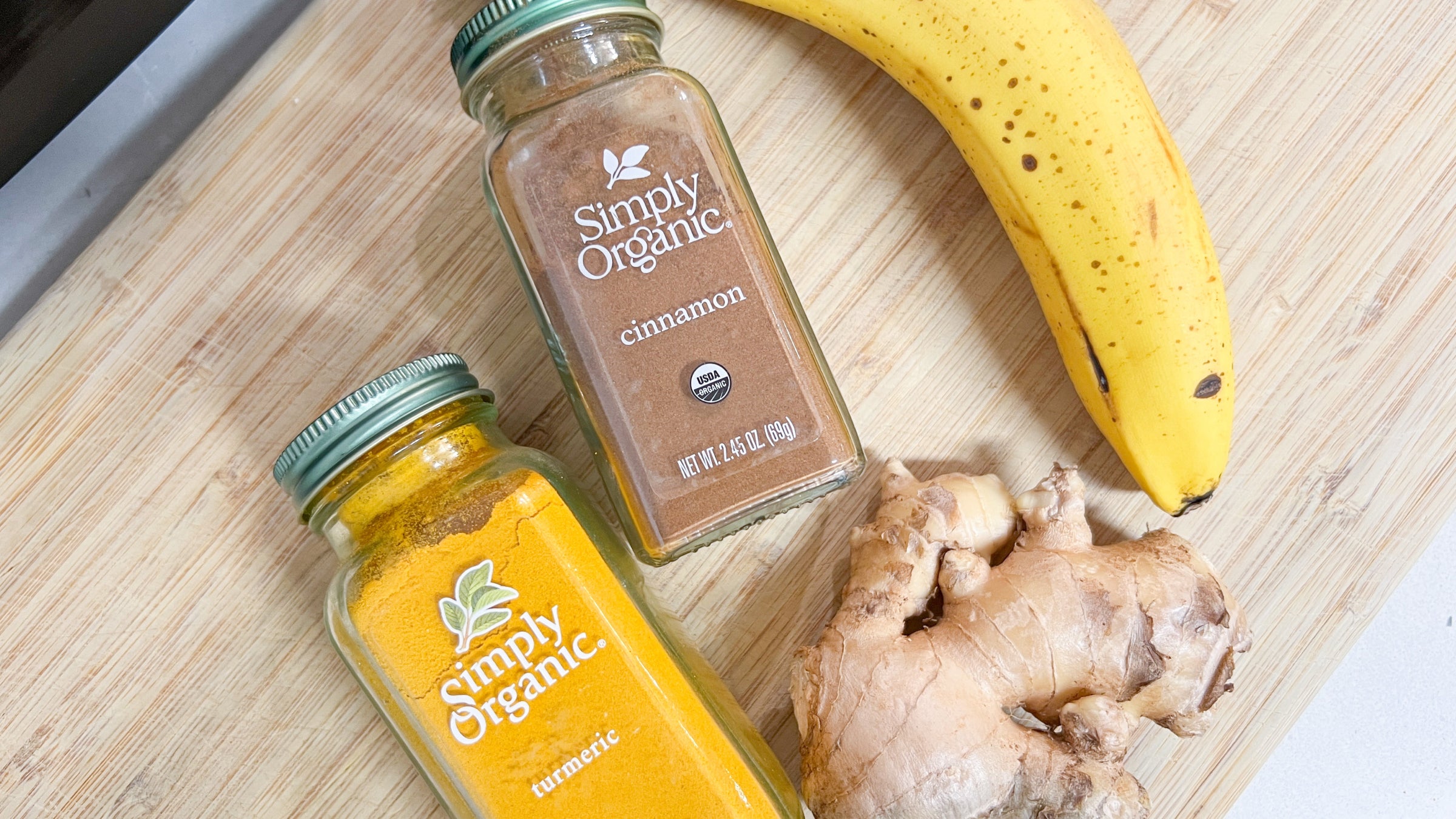 Jars of simply organic turmeric and cinnamon lying on a cutting board with a banana and piece of ginger