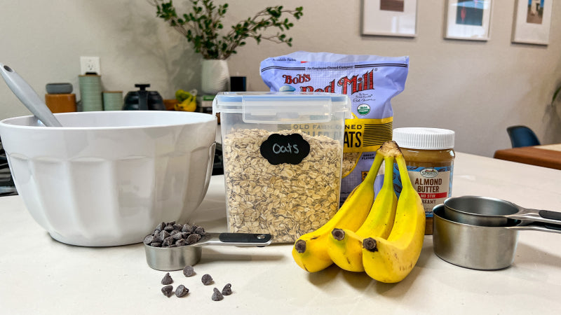 Oats bananas nut butter and chocolate chips sit on the counter next to a white mixing bowl and measuring cups