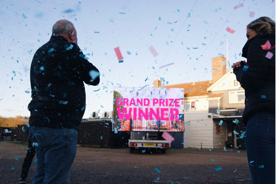Confetti surrounding large TV screen reading 'Grand Prize Winner' with the winner and presenter of the Omaze Million Pound House Draw