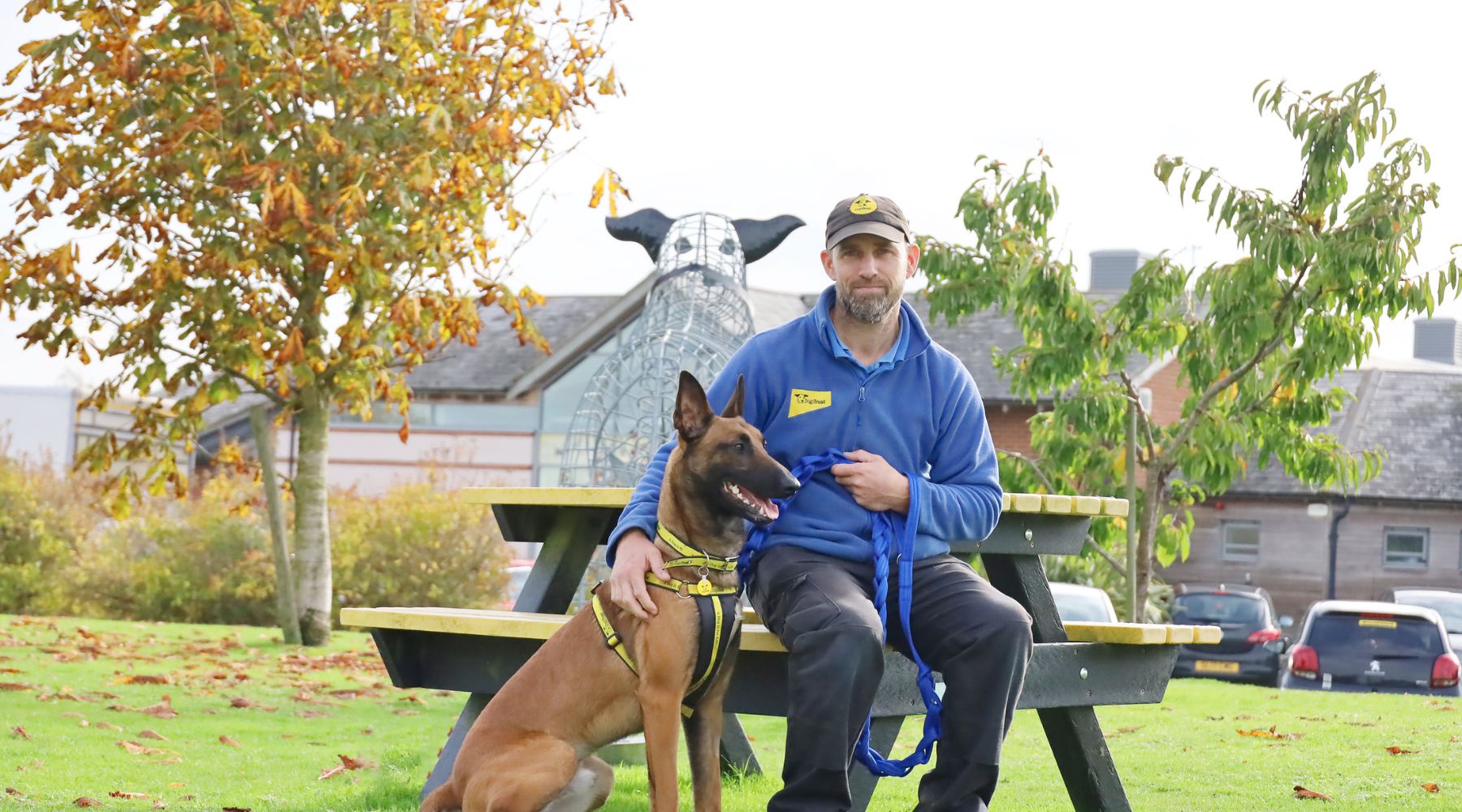 Omaze Million Pound House - Piper the Belgian Malinois sits next to his Dogs Trust trainer on a bench outside the rehoming centre