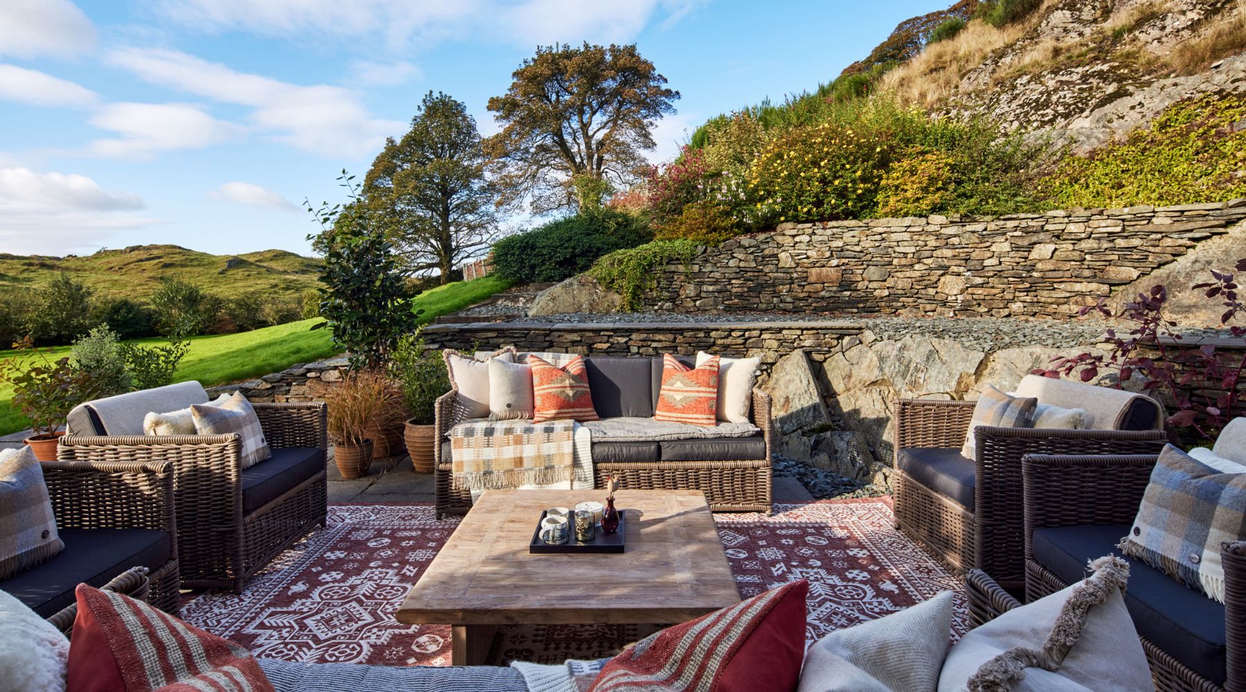 Omaze Million Pound House Lake District  Outdoor lounge with wicker sofas surrounded by drystone walls and mature trees in the sun