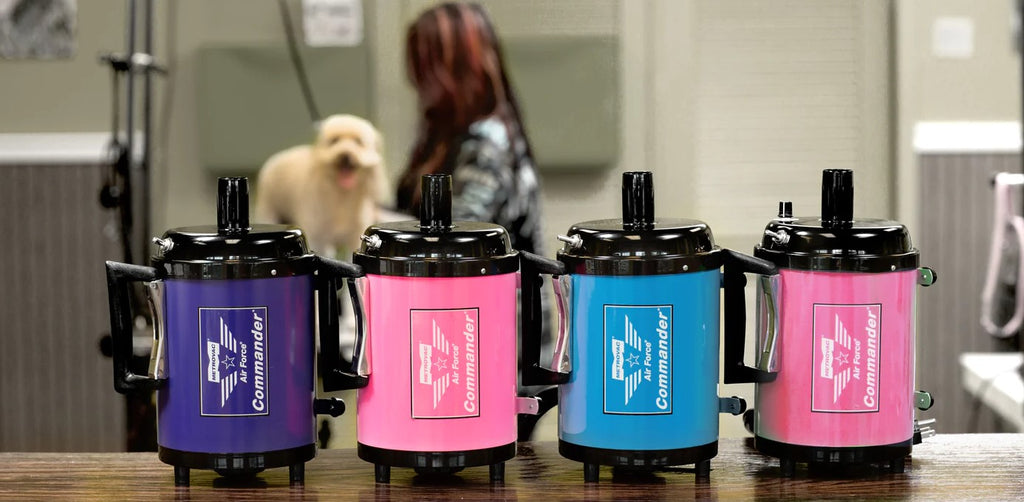 MetroVac Commander Dryers Made in the USA lined up on wood table. Left to right: Purple, pink. blue, pink. In the background a groomer is grooming a medium sized labradoodle