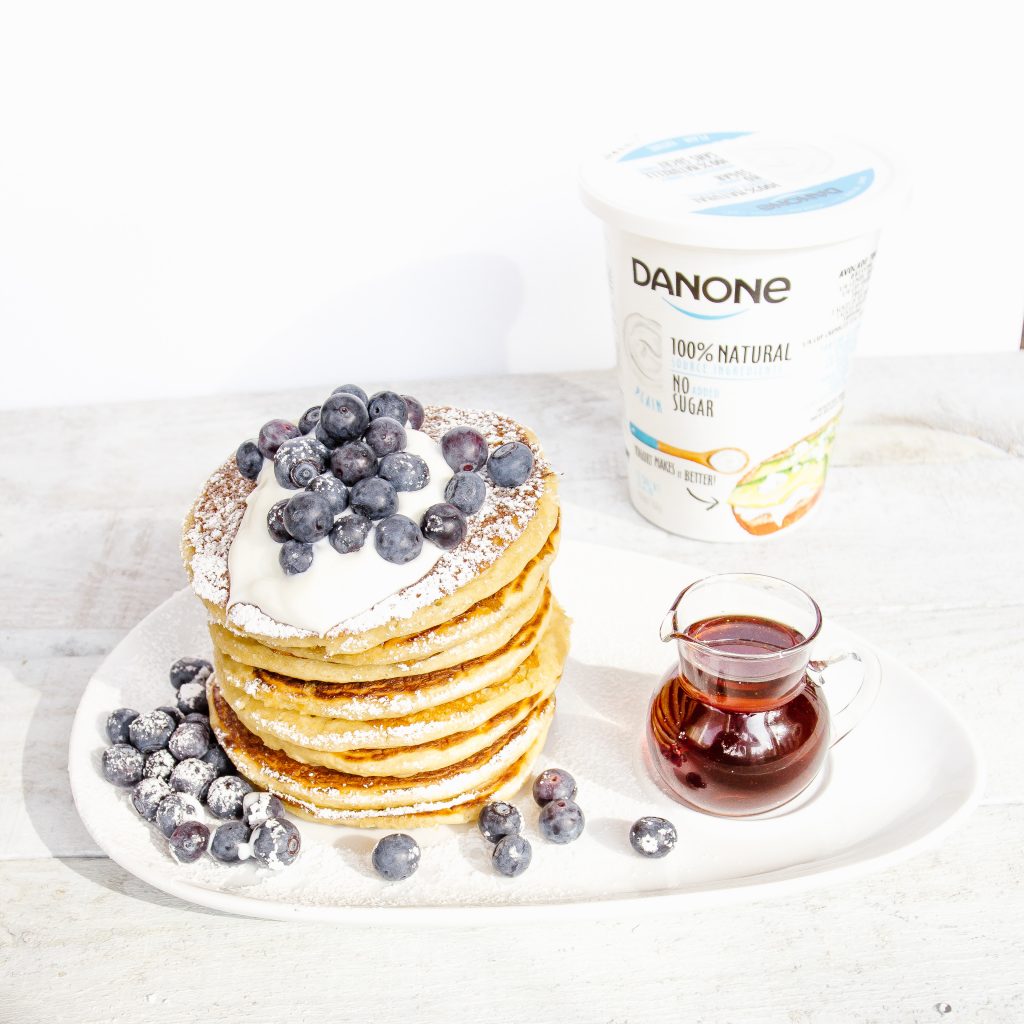 Stack of pancakes topped with Danone plain yogurt and blueberries, with maple syrup and a container of Danone yogurt