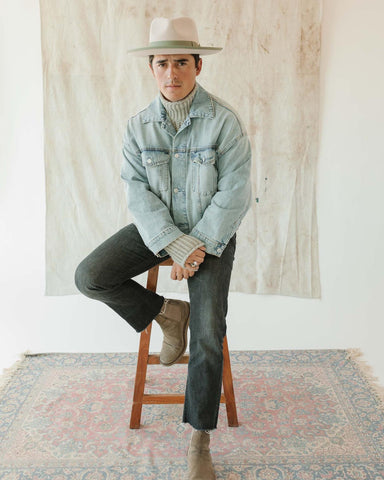 Man sitting on a stool over a patterned rug wearing a denim jacket, black denim jeans, Chelsea boots, and a felt fedora hat