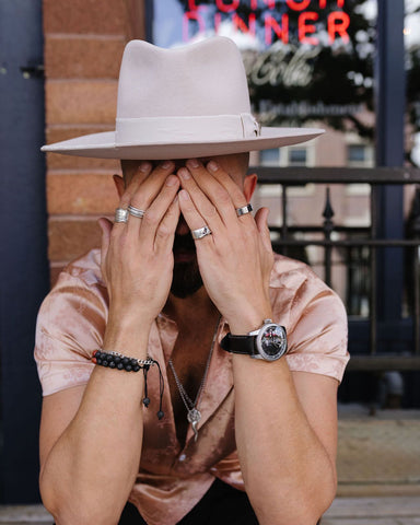 Man covering his face sitting outside of a shop wearing a pink patterned zipper shirt, pink men’s hat, watch, bracelets, and rings.