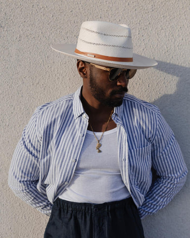 Man leaned up against a concrete wall wearing a pinstriped button-down shirt, necklace, sunglasses, and a white straw hat