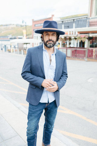 Man on the sidewalk of an empty street wearing a navy suit jacket, button-up shirt, blue jeans, and a navy men’s dress hat