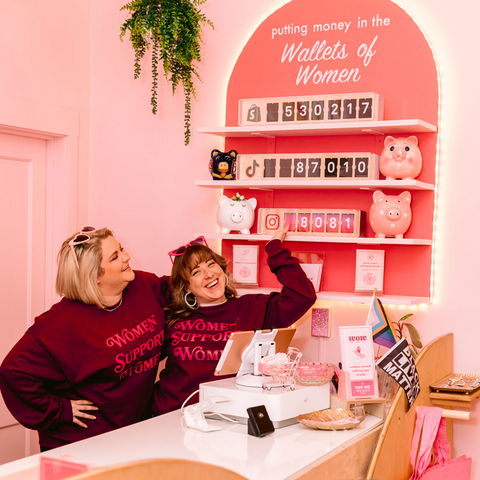 Photo of two women smiling and looking at wall shelves with sales counter and tagline that reads "Putting money in the wallets of women"