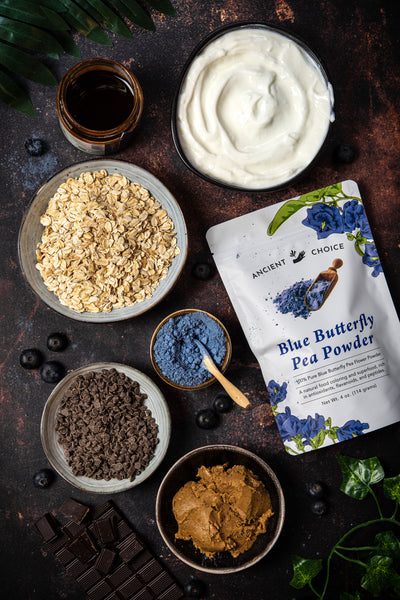 Ingredients on a table: butterfly pea flower powder, nut butter, oats, chocolate, and yogurt