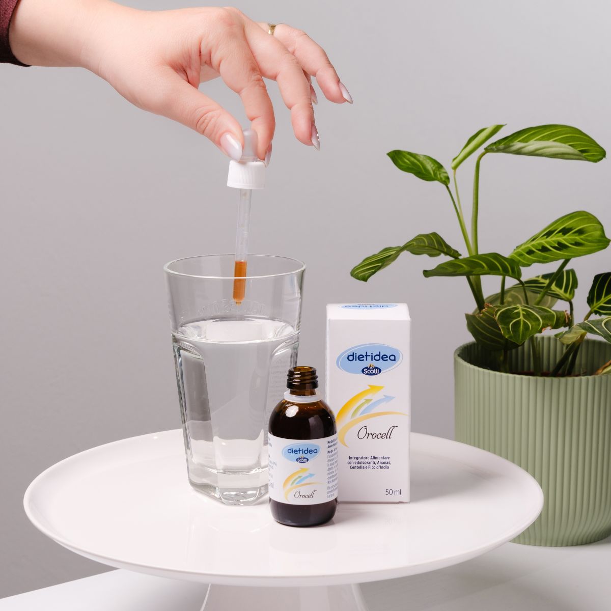 Boccetta di integratore Orocell con a fianco la sua confezione e un bicchiere d'acqua in cui tramite una pipetta viene diluita la soluzione