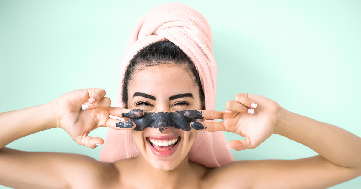 A smiling woman spreading activated charcoal mask on her face.