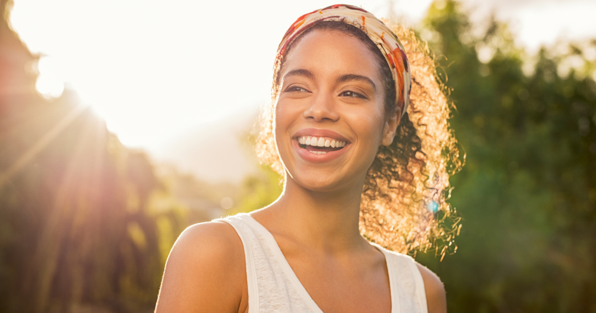 A black woman smiling with radiant skin standing in the setting sun.