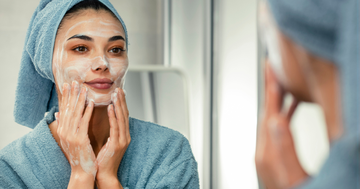 A woman in a gray robe and towel washing her face with tumeric facial cleanser by Goddess of the Sea.