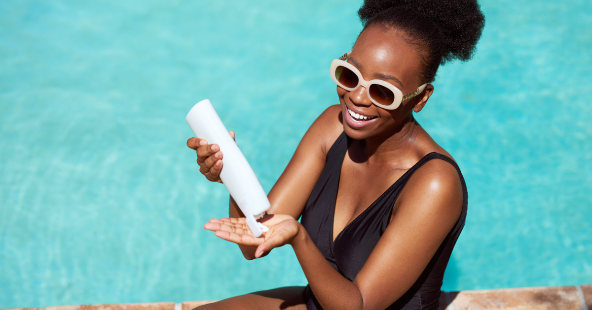 A black woman in a black bathing suit by the pool putting on sunscreen.