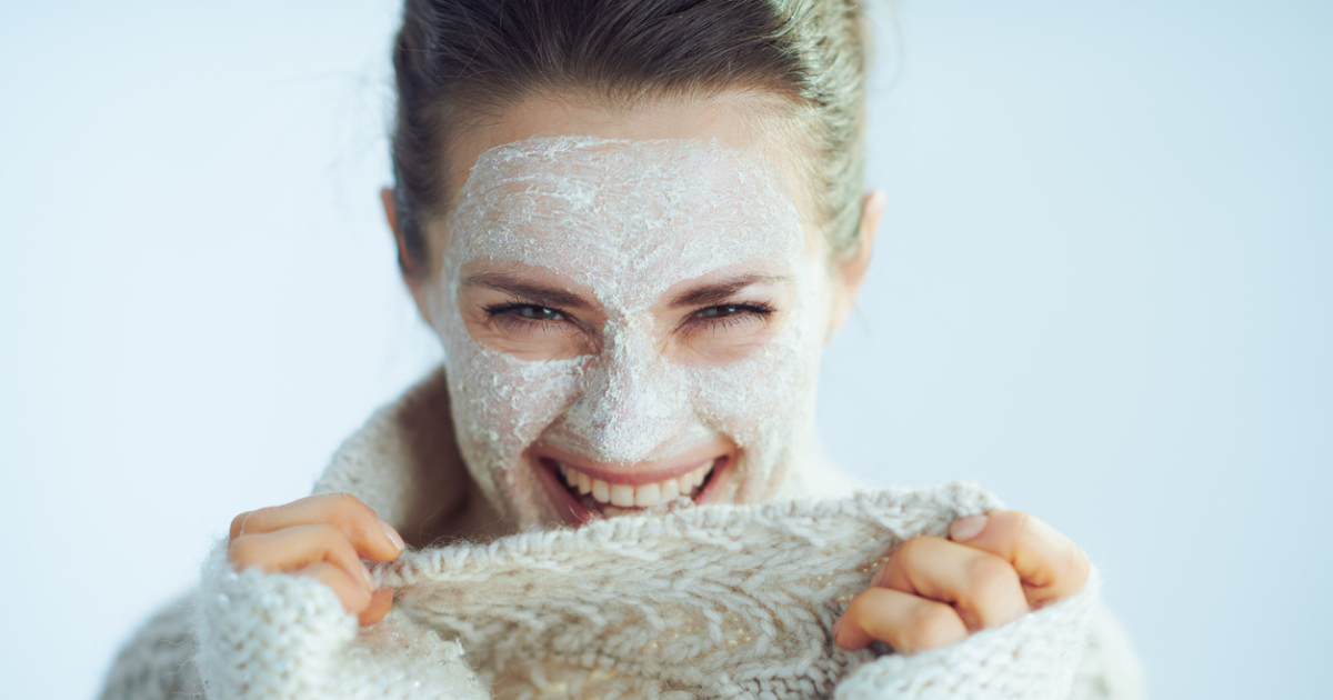 A woman smiling wearing a sweater and applying a face mask.