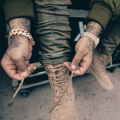 desert boots being tied up by model with gold watch, tattoos and bracelet green stacking trousers an