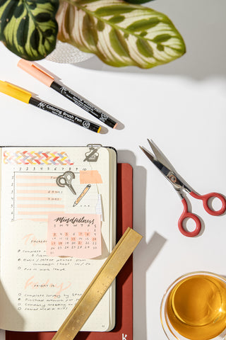 A dot grid notebook lays face-up on the table, its dot grid pages filled with notes and designs. A brass ruler lays across the page, while scissors, markers and a cup of tea lay around the notebook on a white surface