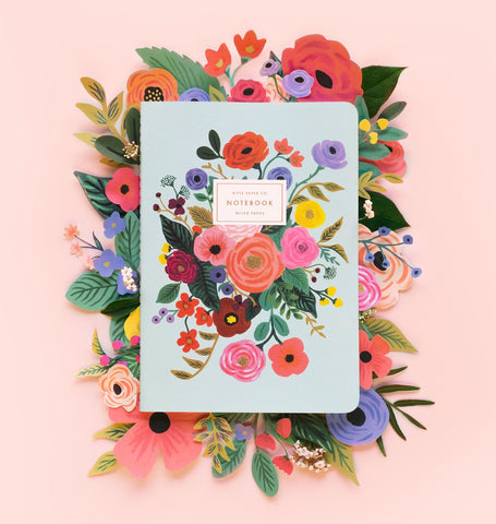 A soft cover Rifle Paper Co. notebook with a floral design on its pale blue cover, on a bed of papercraft flowers in the same style as the notebook art