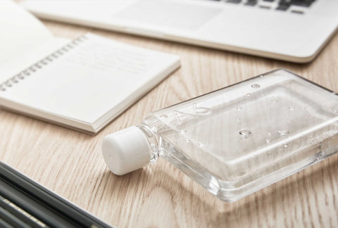 An A6 memobottle (a clear, rectangular bottle) lays flat on a blonde wood desk, with an open notebook and laptop nearby.