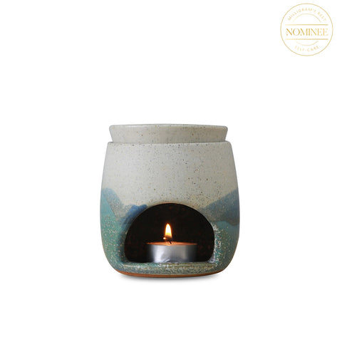 A small cylinder of cream and blue ceramic with rounded edges, with an arch-shaped opening holding a tea light candle