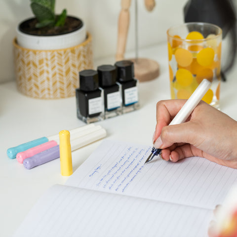 Pilot's Iroshizuku inks next to a handful of colourful Kakuno pens as a person writes on a sheet of paper