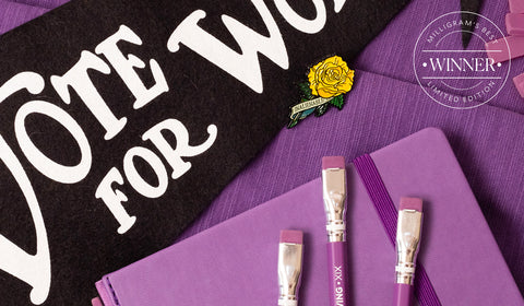 Blackwing's Volume XIX pencils with a purple finish and erasers laid on top of a matching purple notebook next to a black pennant flag with text reading Votes For Women