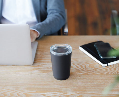 A Kinto To Go Tumbler (black) sits on a light wooden table, with a notebook and laptop nearby