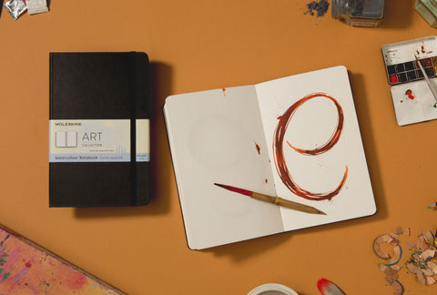 A closed Moleskine Art Watercolour Notebook sits on a bench next to an open version with a vibrant, dramatic 'e' painted in watercolour on one plain page.