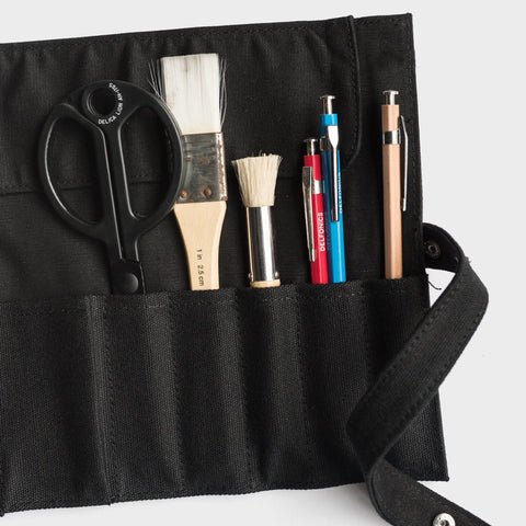 Delfonics Canvas Roll Pencil Case | Milligram [img: a black piece of canvas lays flat on a white surface, with pens, brushes and scissors held in its slim vertical pockets. A strip of canvas with a push-stud hangs off one side of the canvas piece]
