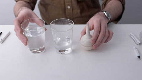 Two glasses of water sat on a white table beside a bulb syringe (a squeezy ball with a narrow opening used to puff air or squirt water in a controlled manner)