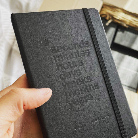 A black Moleskine notebook with "10 seconds minutes hours days weeks years" embossed on the front