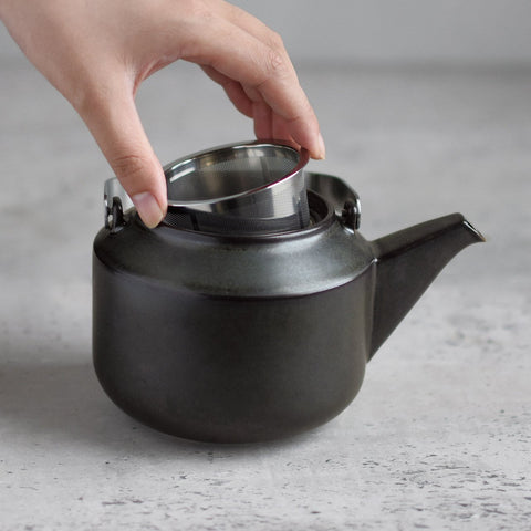 Kinto's Leaves To Tea teapot in a dark ceramic finish with a hand holding the stainless steel filter over it